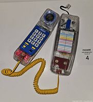 Image showing the full vintage transparent acrylic touch phone with handset and base, coiled yellow cord connecting the two parts, and visible keypad with blue panel and rotary dial style part.