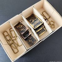 Box showing three hammered copper buckles, three brass buckles and thirteen loop closures