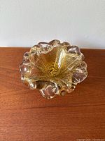 Top view of the clear glass bowl with a wavy, ruffled edge and embedded gold flecks inside, placed on a wood surface against a white wall.