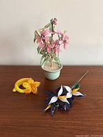 Image showing three glass flower items on a wooden surface against a plain wall: a potted pink flower plant, a yellow daffodil decoration, and a blue and white calla lily bouquet.