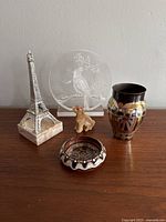 Front view of five items: metal Eiffel Tower on marble base, etched glass bird plate on stand, small brown dog figurine, patterned ashtray, and decorative metal vase.