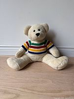 Front view of the beige teddy bear wearing a multi-colored striped knit sweater, sitting on the floor.