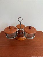 Front view of the condiment holder showing 3 glass dishes with teak lids on a teak base with a central metal ring handle.