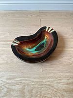 Top-down view showing the kidney-shaped ceramic ashtray with organic glaze of brown, black, and green tones on a light wooden floor.