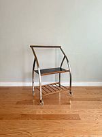 Front angle view of the mid-century bar cart showing its metal frame, wooden top and bottom slatted shelves with castor wheels on hardwood floor.