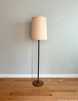 Full view of the mid-century floor lamp showing the lamp with cream fabric shade and bronze metal base standing on wooden floor against white wall.
