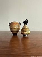 Front view of both miniature Greek ceramic vessels on wooden surface against plain wall.