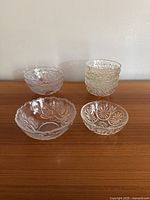 Front view of the eight clear glass serving bowls arranged on a wooden surface against a plain wall. Four stacked smaller bowls on the right and four larger bowls spaced around on the left.