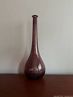 Full view of the tall vintage purple glass vase showing its elongated neck and bulbous base on a flat surface against a plain background.