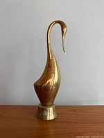 Full front view of vintage brass swan statue on wood surface against plain wall, showing overall form and base with slight wear.
