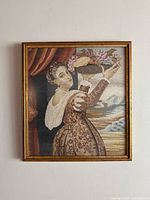Full view of framed vintage needlepoint artwork showing a woman in period dress holding flowers against a cloudy sky background.