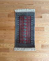 Top-down image of a small rectangular vintage rug laid flat on a wood floor, showing central red patterned field and fringed edges.