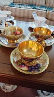 Three tea cup and saucer sets on wooden table; two fruit motif sets with gilt interiors at front and a rose floral set at back