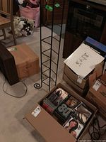 Photo showing box of assorted CDs near a slender tall black metal CD rack on carpeted floor. Background includes boxes and storage items.