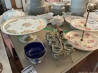 Wide view of table showing array of stacked plates in floral and gold leaf patterns, cobalt blue glass container in silver holder, vintage green glass condiment holders with gold trim, and two-tier floral cake stand.