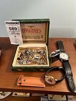 Wide view showing Longines presentation box filled with assorted cufflinks and tie pins on wooden surface next to two wristwatches and leather case