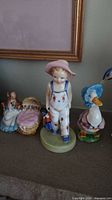 Four figurines displayed together: Jemima Puddleduck, boy with pink hat and dog, Royal Doulton Golliwog, and Hunca Munca.
