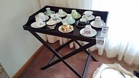 Black wooden folding snack table with crossed legs and rectangular tray top, shown with teacups but contents not included.