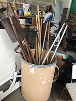 Various long-handled garden tools stored upright in a beige bucket, showing handles, metal heads, broom, and ski pole.