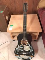 Full-front view of black acoustic guitar with Hawaiian stencil artwork showing palm trees and tropical leaves on body and fretboard visible