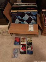 Box of vinyl LP albums inside cardboard box, showing The Cars 'Panorama' at front, with multiple other albums behind. Below box, several cassette tapes arranged on carpet including U2, The Doors and others.