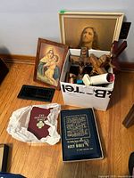 Overview of lot on hardwood floor showing box of crucifixes, rosaries, Bibles and framed prints