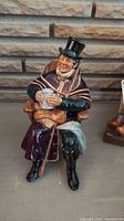Front and angled front view of the Royal Doulton The Coachman figurine holding a mug, seated on a chair with detailed clothing and facial expression.