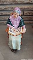 Front view of seated elderly woman figurine looking at photo album, detailed facial features and clothing colors.
