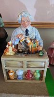 Front view of Royal Doulton figurine The China Repairer showing seated elderly man at table with china dolls and tools.