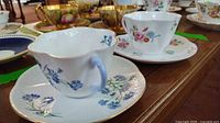 Two Shelley fine bone china teacups and saucers placed on a wooden table, one with blue cornflower design and the other with multicolor floral design, both with gold trim.