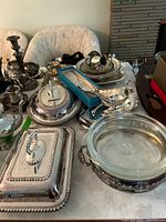 Table showing assortment of silver plated serving dishes, covered trays, tea set, utensils, and gravy boat with glass insert.