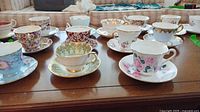 Wide view of 10 assorted teacups and saucers with floral and gilt gold patterns on wooden table.