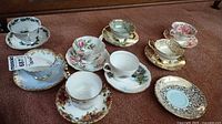 Photo showing six assorted vintage tea cups and saucers arranged on carpet, showcasing varied floral patterns and gold accents