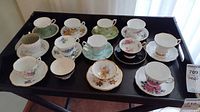 Wide view of all 11 teacup and saucer sets arranged on black tray showing varying floral patterns, colors, and gold accents.