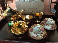 Full set of Royal Chelsea gold tone teapot, creamer, bowl, and cup with saucer along with two floral ceramic cups and saucers displayed on a dark surface.