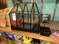 Glass terrarium with black metal frame and pitched roof design shown on wooden shelf, items inside visible.