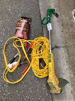 Green weed trimmer, yellow extension cord coiled, and booster cables with red and black clamps lying on ground