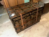 Closed view of vintage steamer trunk showing wood slats, metal hinges, corner reinforcements and leather straps.