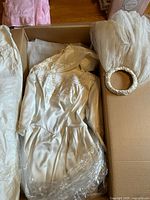 Box containing the vintage wedding dress folded, showing the skirt, bodice, and crinoline fabric along with the headpiece and veil beside it.