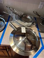 Stack of five stainless steel frying pans with one lid on top and MaxSold tag visible
