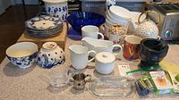 Wide view of countertop displaying multiple ceramic plates and bowls, gravy boat, mugs, mortar and pestle, butter dish, cookie jar, and other kitchen accessories.
