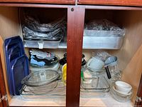Overall contents of the cabinet showing all items in the lot including blue pans, clear Pyrex glass bakeware, metal pans, glass and ceramic bowls, measuring cups, and gloves.