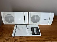 Pair of white Bose Model 100 speakers shown side by side on wooden surface with user manual in front.