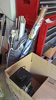 Photo shows metal and plastic tubular parts for canopy frame partly inside a grey bag, along with one packaged blue and white umbrella with wooden handle and a black metal and glass lantern inside a box.