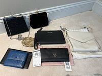 Photo showing six items on a carpeted floor including black beaded clutch, small gold coin purse, black wristlet, blue and black bi-fold wallet, white crossbody bag, and black Radley wallet.