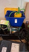 Blue storage bin containing a yellow and green spray pump, assorted sprayers, and garden chemicals. Boxes containing a 25ft Rapid Flow hose and Melnor Turbo Oscillator sprinkler visible underneath and around bin.