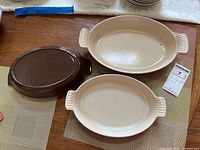 Three Le Creuset oval ceramic bakeware dishes shown from top and side view, two ivory and one dark brown, placed on table mats.