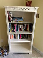Front-facing image of white wood Lexington bookcase filled with books on three adjustable shelves. Shows the decorative cutout pattern on top panel and fluted sides.