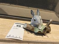 Porcelain figurine of a white and gray bunny on a brown branch with green leaves and white flowers. Some leaves are broken.