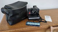 Photo showing black zipped Polaroid branded carrying case, Polaroid Pronto RF camera, flash bar, and camera manual all on wooden surface.